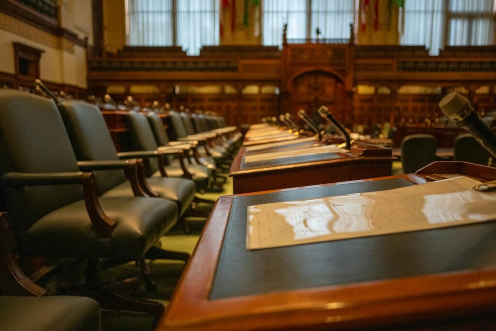 A close-up view of empty parliamentary or legislative chamber seats and desks, each equipped with microphones and documents for ESG Reporting. The room features traditional wood-paneled walls, high-backed chairs, and large windows with vertical blinds, suggesting a formal governmental or institutional setting.