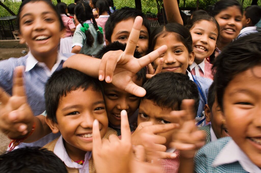 A group of joyful schoolchildren in uniforms smiling and flashing peace signs while standing closely together outdoors, capturing a moment of playful energy and friendship.