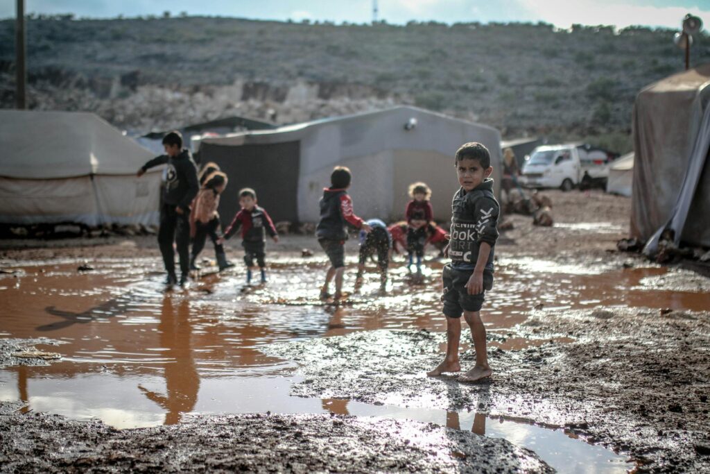 Children playing barefoot in muddy puddles at a refugee or displacement camp surrounded by tents and makeshift shelters, reflecting harsh living conditions and poverty.