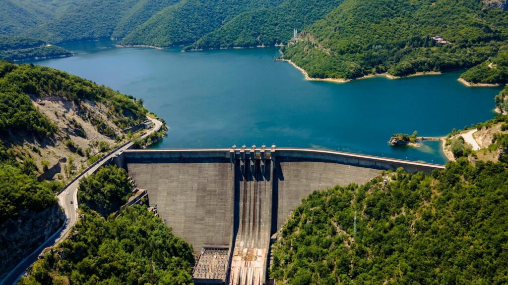 Aerial view of a large hydroelectric dam nestled between forested mountains, holding back a vast, clear blue reservoir. The infrastructure symbolizes regional water management. This image represents Transboundary Water Cooperation, highlighting the legal and diplomatic frameworks needed to manage shared water resources between nations peacefully.