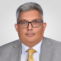 Head-and-shoulders portrait of a man with gray hair and glasses wearing a gray suit and yellow tie against a neutral backdrop.